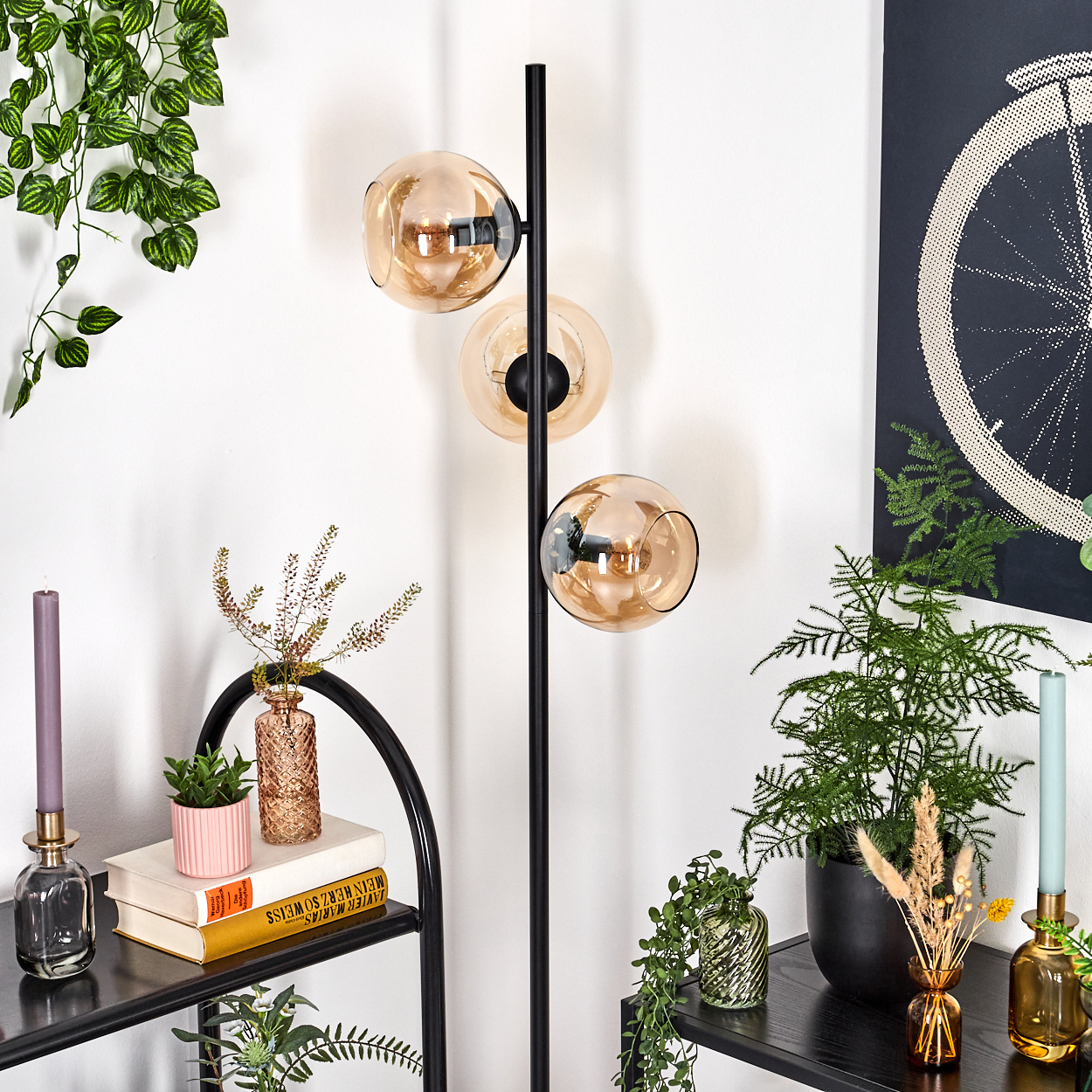 Ein modernes Zimmer mit einer schwarzen Stehlampe mit drei Kugelleuchten, Pflanzen und Dekorationsartikeln auf Regalen. Ein Fahrradrad-Kunstwerk ist an der Wand.
