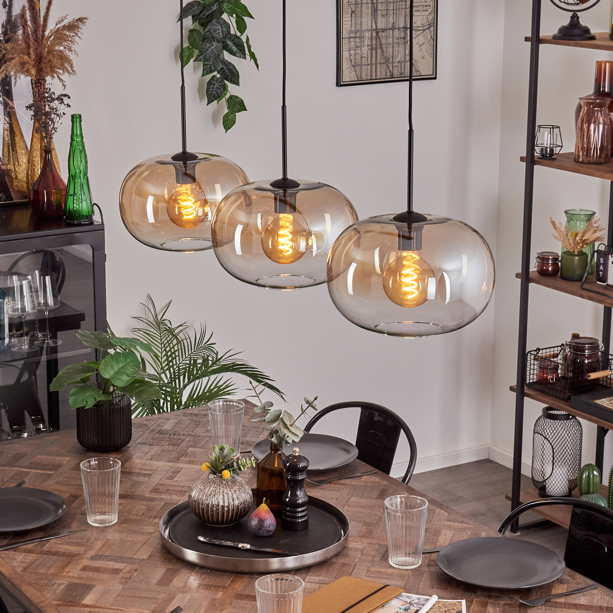 Esszimmer mit Holztisch, drei Hängelampen und Dekorationen. Teller, Gläser und ein Tablett mit Obst stehen auf dem Tisch.