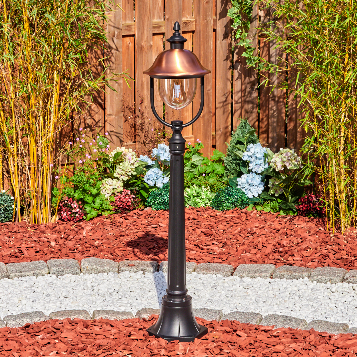 Eine schwarze Lampe mit kupferfarbenem Dach steht in einem Garten. Bambus und Blumen umgeben sie. Roter Mulch und Stein säumen den Weg.