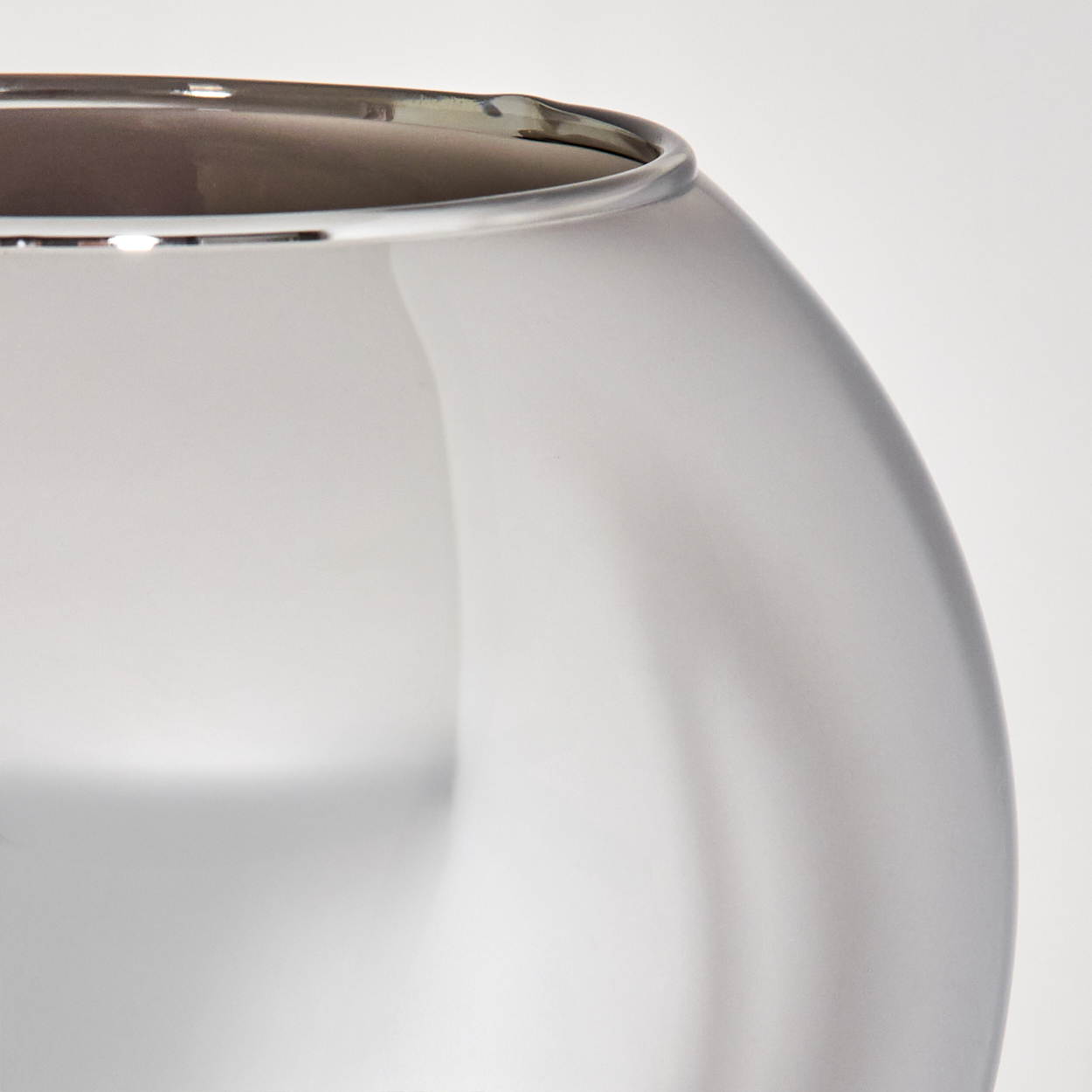 Nahaufnahme einer Vase aus mattiertem Glas mit silbernem Rand, vor weißem Hintergrund. Das Innere der Vase ist dunkel.
