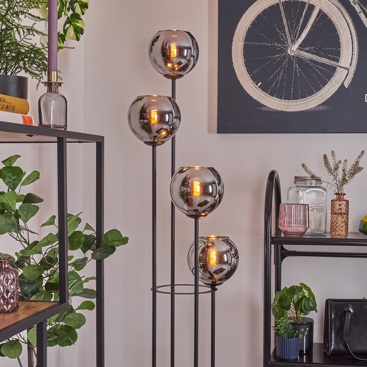 Eine moderne Stehlampe mit vier Glaskugelschirmen ist der Mittelpunkt. Regale zeigen Pflanzen und Dekoration, Fahrradkunst an der Wand.