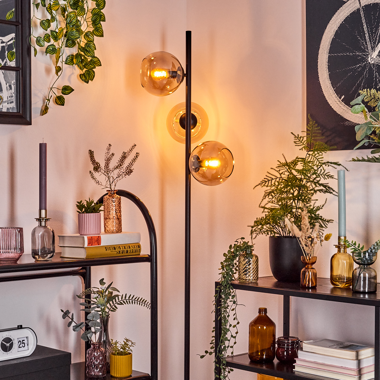 Eine Zimmerecke mit einer Lampe mit drei Glühbirnen, Regalen mit Pflanzen und Dekoration sowie Wandkunst. Eine Kerze und Bücher sind zu sehen.