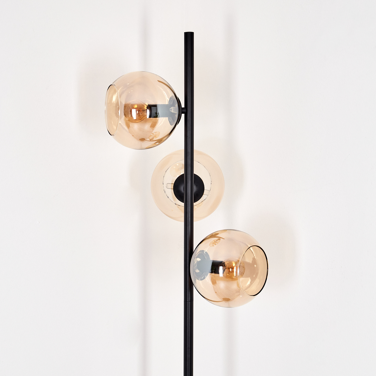 Eine moderne Stehlampe mit drei kugelförmigen, bernsteinfarbenen Glasschirmen. Die Lampe hat ein schwarzes Metallgestell, vor einer weißen Wand.