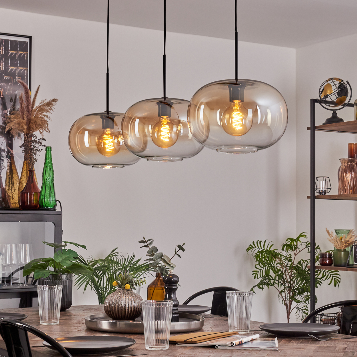 Esszimmer mit drei Hängelampen. Holztisch mit Tellern und Gläsern. Pflanzen und Dekorationen sind sichtbar.