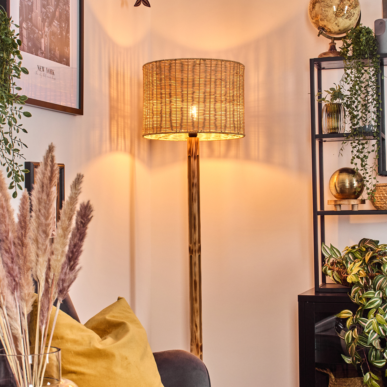 Eine beleuchtete Korblampe steht in einer Ecke. Dahinter ein Regal mit Globus und Pflanzen. Davor eine Vase mit Pampasgras.