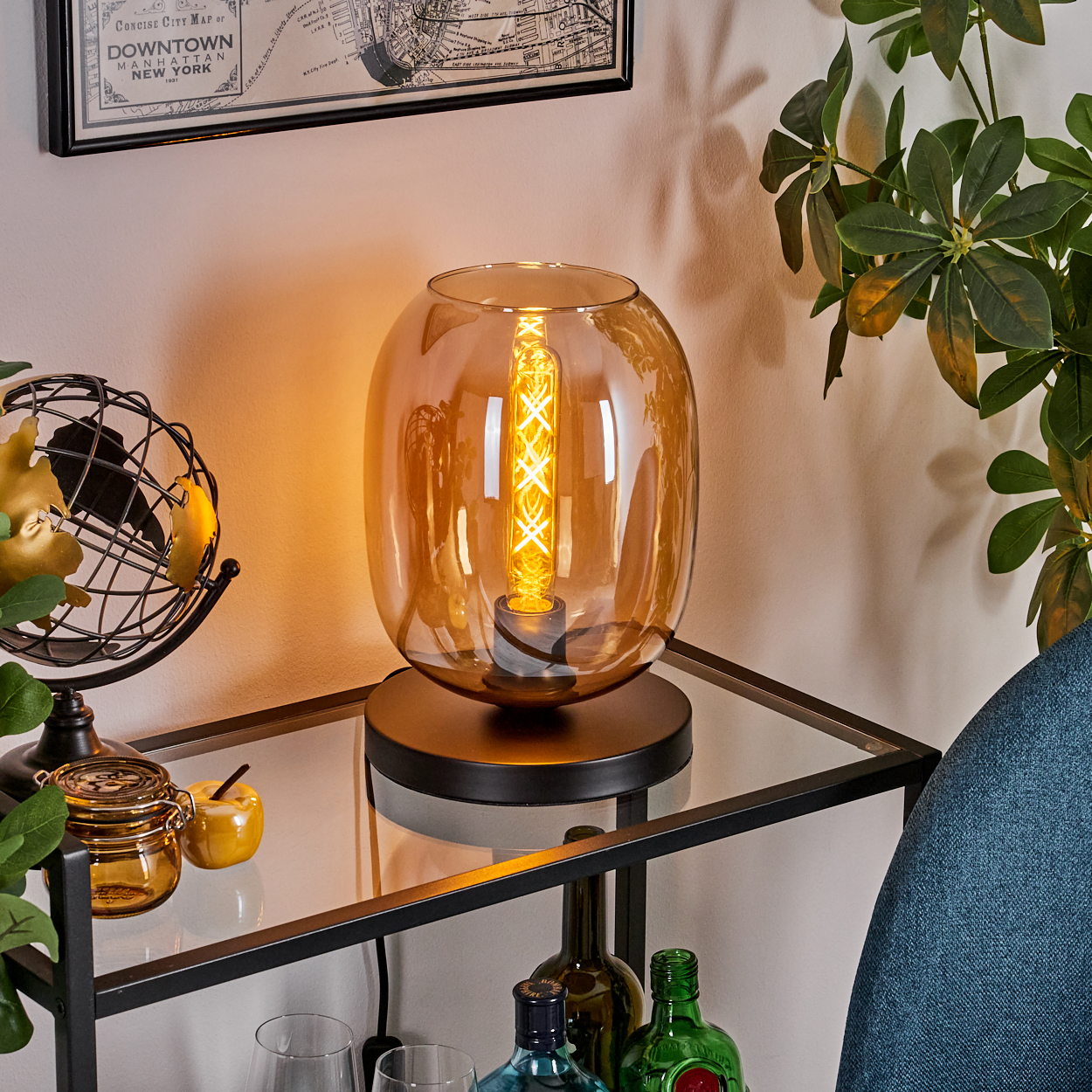 Eine beleuchtete, bernsteinfarbene Glaslampe steht auf einem Glasregal. Globus, Glas und Flaschen sind in der Nähe.