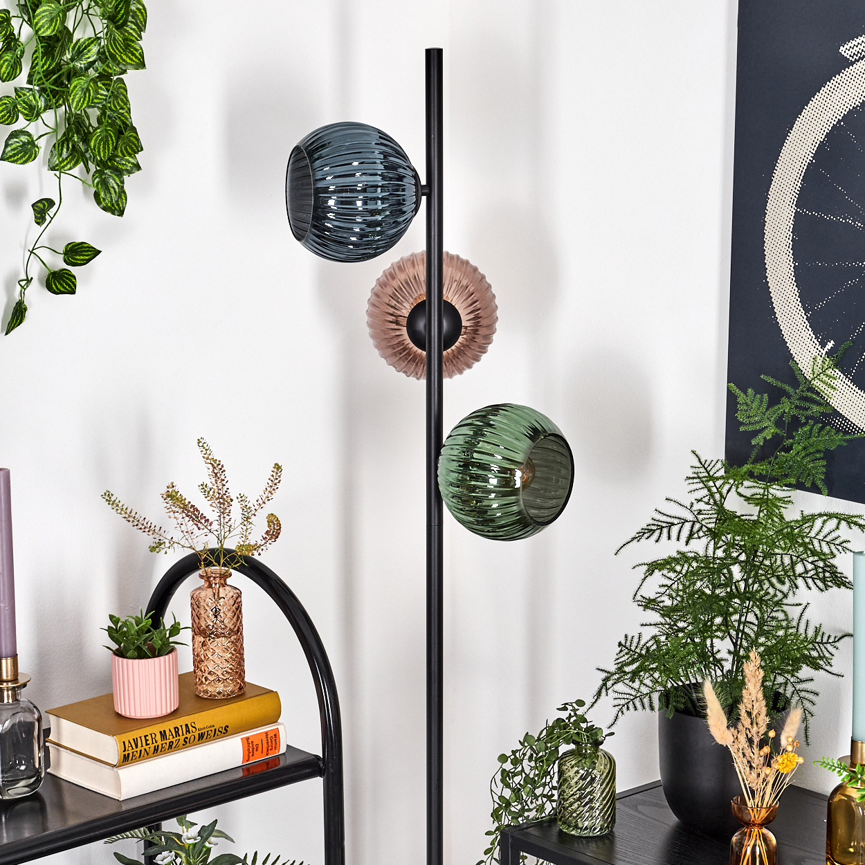 Eine moderne Lampe mit drei Glasschirmen in verschiedenen Farben. Pflanzen und Bücher im Vordergrund, ein Fahrradrad-Kunstwerk im Hintergrund.