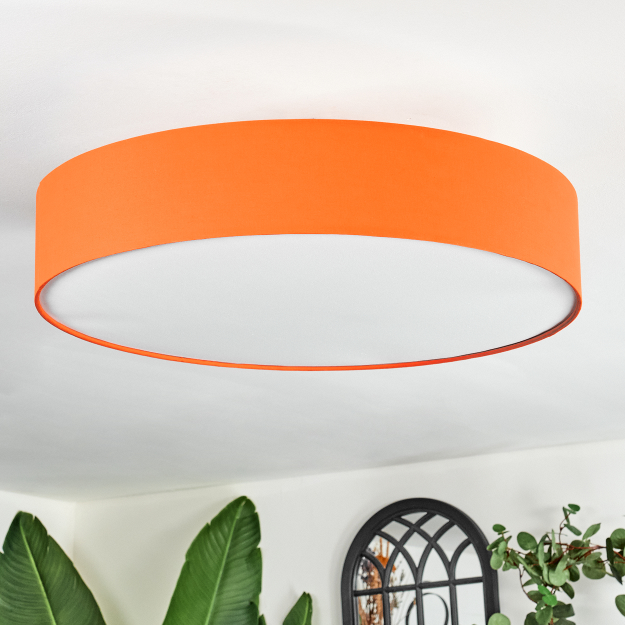 Ein orangefarbener Lampenschirm hängt von der Decke. Darunter sind eine grüne Pflanze und ein dekoratives schwarzes Fenster zu sehen.