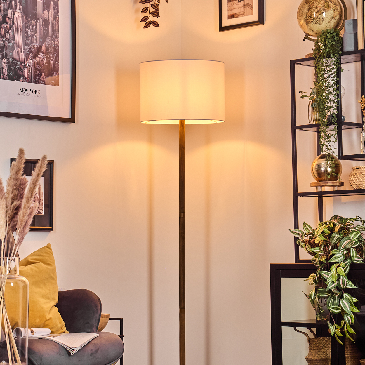 Eine Stehlampe steht in einem Raum mit Regal, Sessel, Bildern und Pflanzen. Warme Beleuchtung erhellt die Szene.