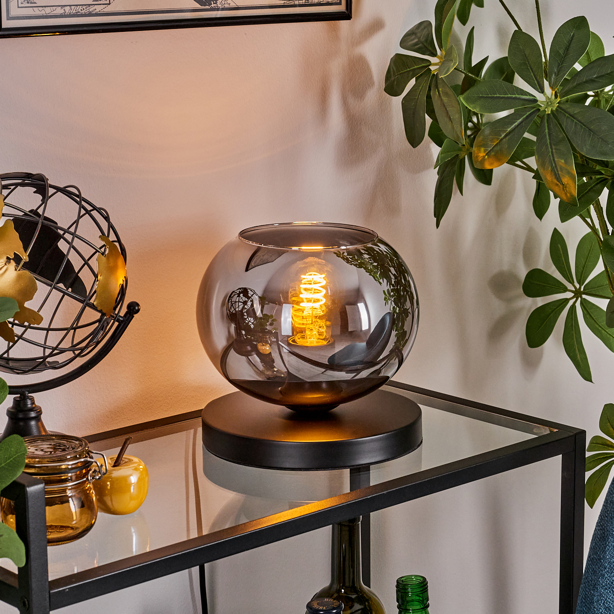 Eine dekorative Lampe mit leuchtender Glühbirne steht auf einem Glasregal. Ein goldener Globus und Pflanzen sind ebenfalls zu sehen.