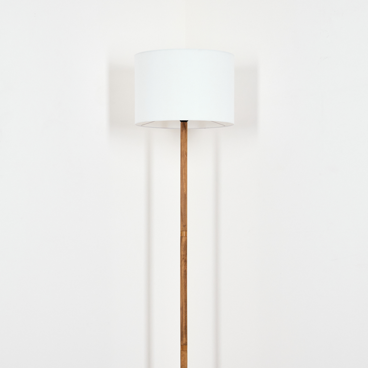 Eine Stehlampe mit weißem Lampenschirm und Holzstange steht vor einer weißen Wand. Die Lampe wirft einen Schatten an die Wand.