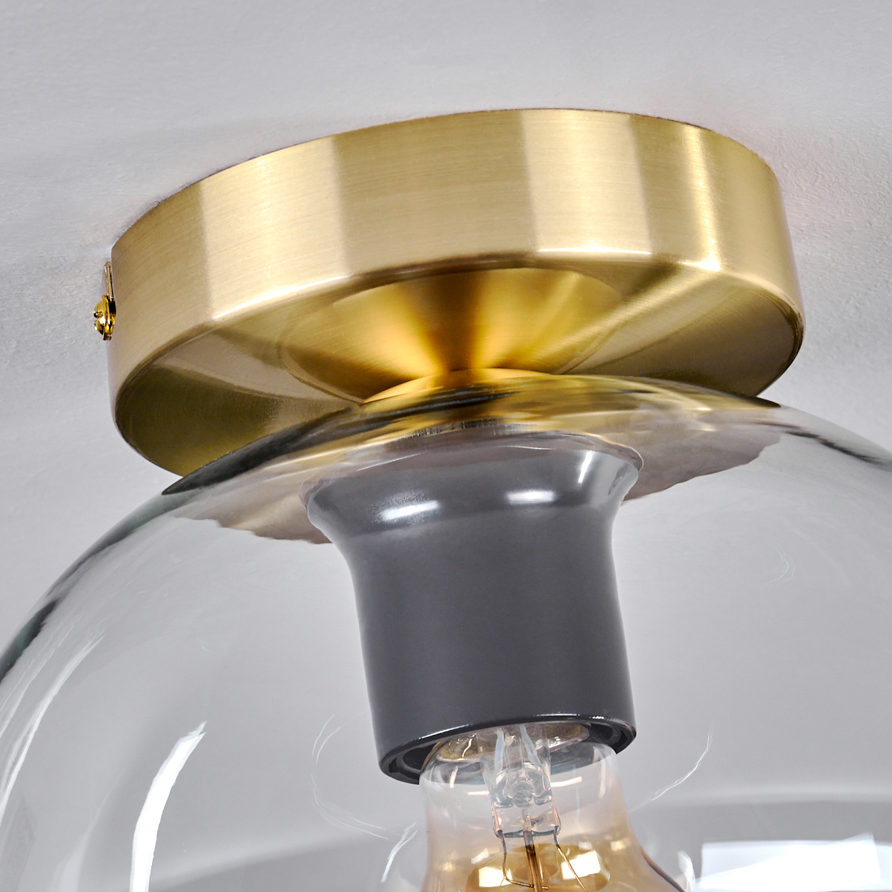 Nahaufnahme einer Deckenleuchte. Sie hat einen goldfarbenen Sockel, einen klaren Glasschirm und eine sichtbare Glühbirne.
