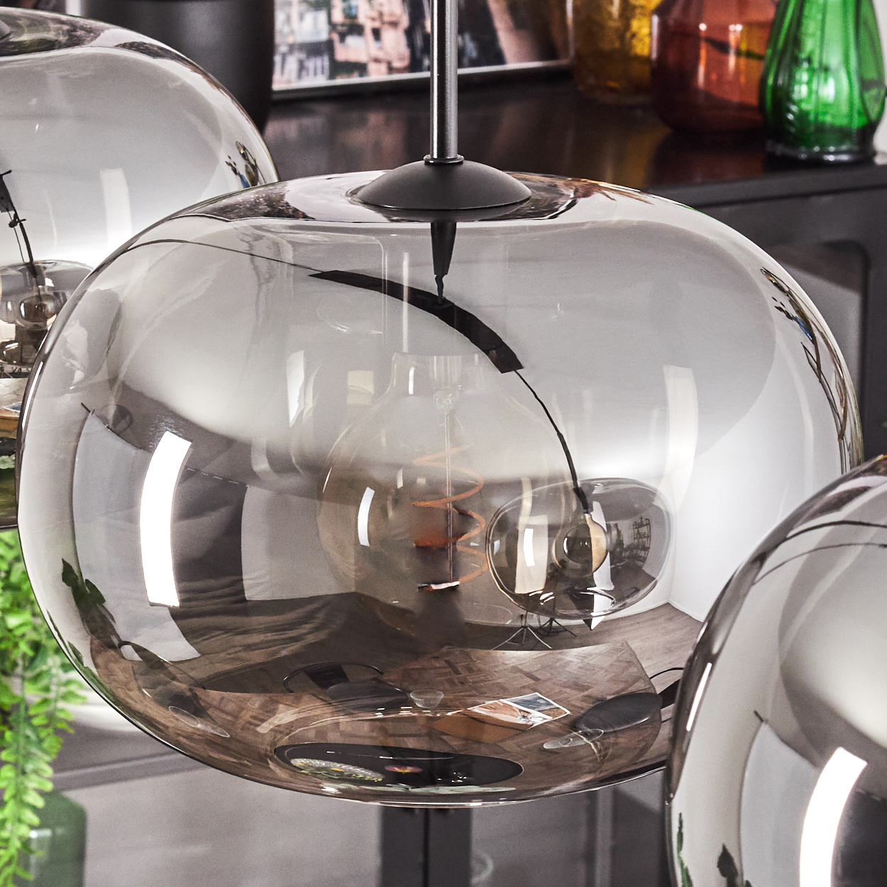 Nahaufnahme von modernen Hängeleuchten. Jede Leuchte ist eine große, rauchige Glaskugel, die einen Raum mit Holzböden reflektiert. Der Hintergrund umfasst Vasen.