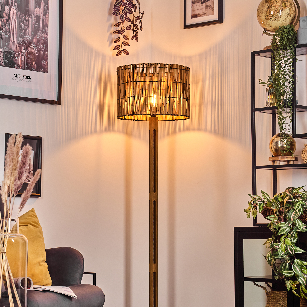 Eine beleuchtete Lampe mit gewebtem Schirm steht in einem Raum mit Pflanzen, einem Globus und gerahmten Bildern. Im Vordergrund befindet sich ein Sofa.