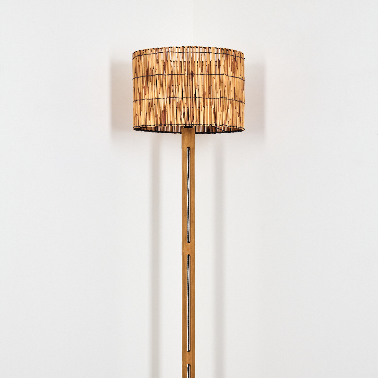 Eine hohe Stehlampe mit Bambusschirm und Holzständer steht vor einer weißen Wand. Die Lampe ist beleuchtet.