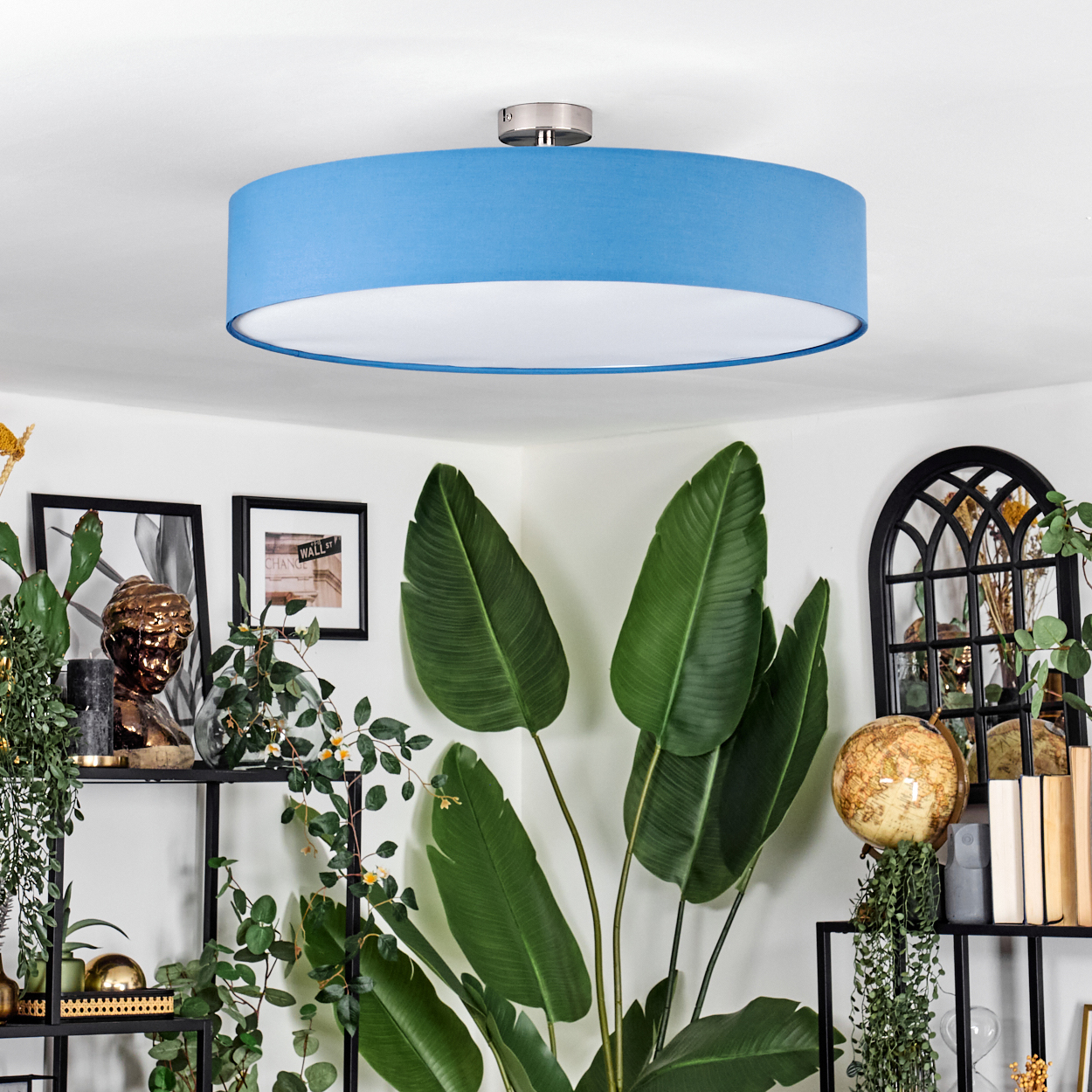 Ein Raum mit blauem Lampenschirm an der Decke. Es gibt Pflanzen, Regale mit Dekorationen und einen Globus. Die Wände sind weiß.