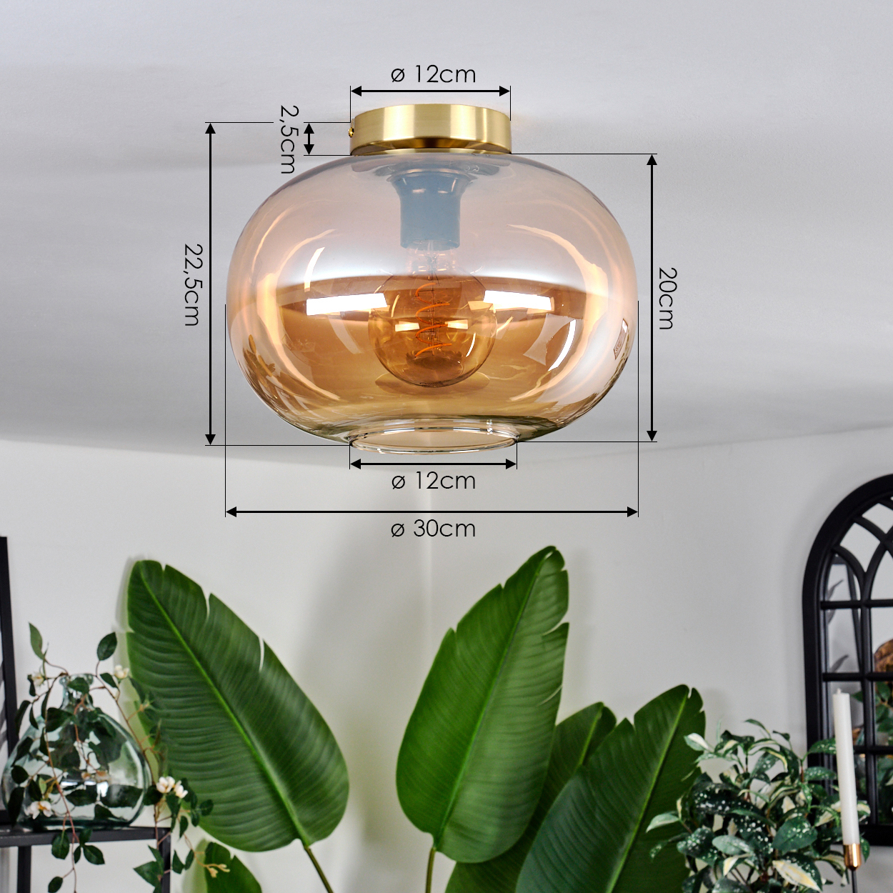 Eine runde Deckenlampe aus bernsteinfarbenem Glas mit goldenem Sockel. Maße werden angezeigt. Große grüne Blätter im Vordergrund.