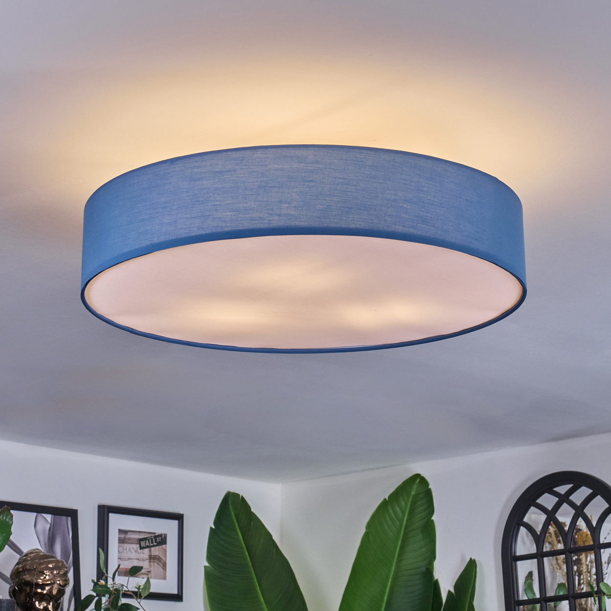 Eine blaue, runde Deckenlampe ist an einer weißen Decke montiert. Darunter sind grüne Pflanzen und gerahmte Bilder zu sehen.