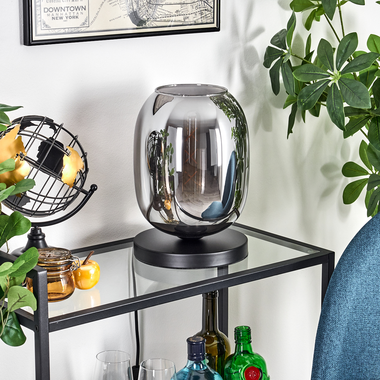 Eine dekorative Szene mit einer reflektierenden Lampe, einem Globus und Pflanzen auf einem Glastisch. Flaschen und eine Karte im Hintergrund.