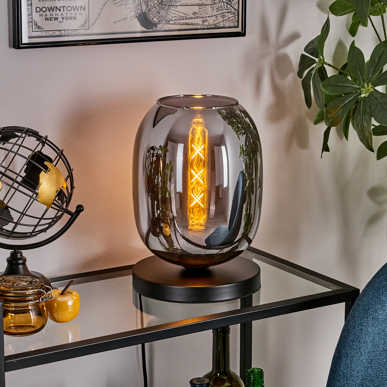 Eine moderne Lampe mit leuchtender Glühbirne steht auf einem Glastisch. Globus und kleine Gläser stehen auf dem Tisch. Karte und Pflanze sichtbar.