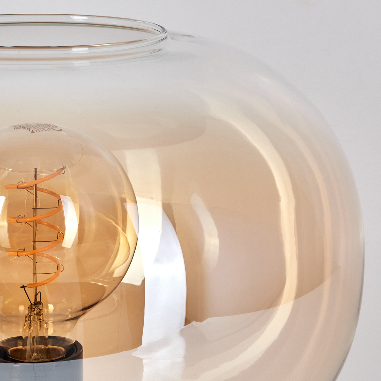 Nahaufnahme einer dekorativen Lampe. Eine goldene Glühbirne befindet sich in einem großen, transparenten, kugelförmigen Glas. Der Hintergrund ist weiß.