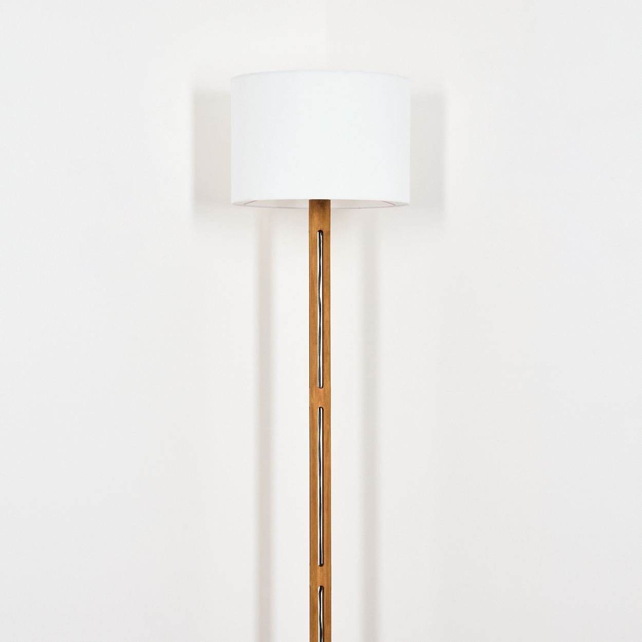 Eine moderne Stehlampe mit weißem Lampenschirm und Holzstange steht vor einer weißen Wand. Die Lampe ist zentriert.