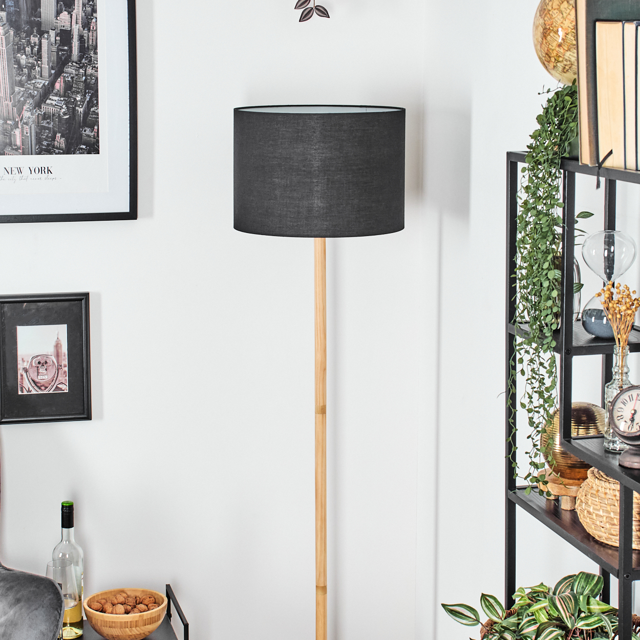Eine Stehlampe mit schwarzem Schirm steht in einem Raum. Regale mit Pflanzen und Dekoration befinden sich rechts, Bilder links.