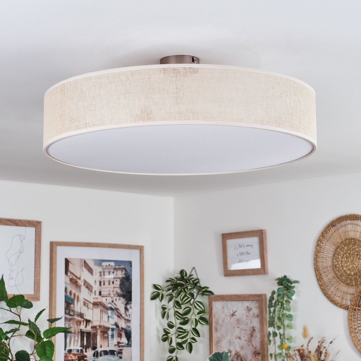 Eine beige Trommel-Deckenleuchte ist zentriert. Darunter befinden sich gerahmte Bilder, Pflanzen und eine Wandbehang aus Weide, alles vor einer weißen Wand.