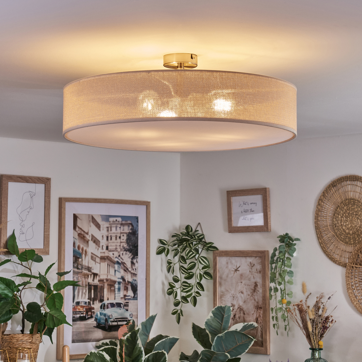 Eine beige Trommeldeckenleuchte beleuchtet einen Raum mit gerahmten Bildern und Pflanzen. Ein Straßenbild ist zu sehen.
