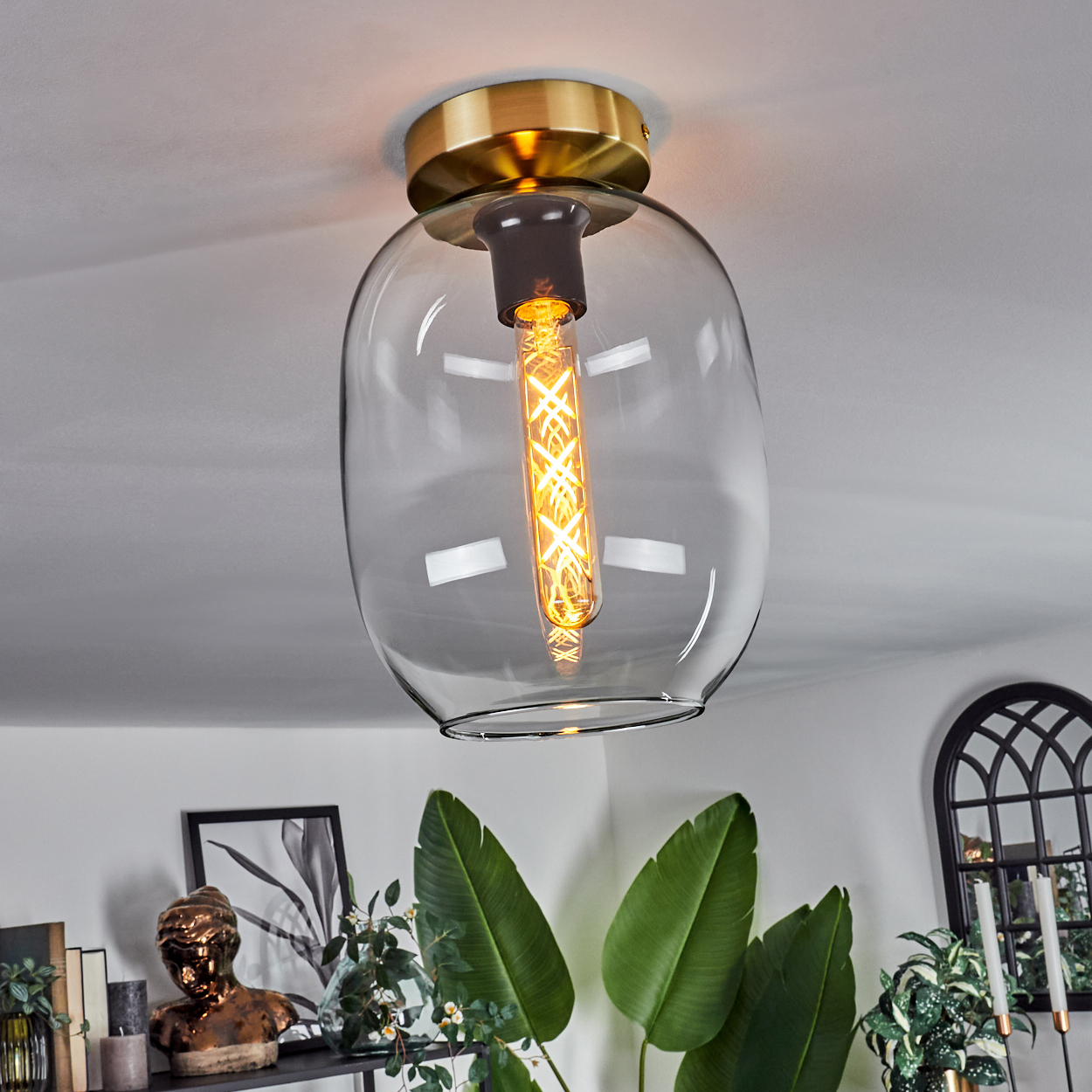Eine moderne Deckenlampe mit klarem Glasschirm und warmer Glühbirne. Grüne Pflanzen und eine Büste sind im Hintergrund.