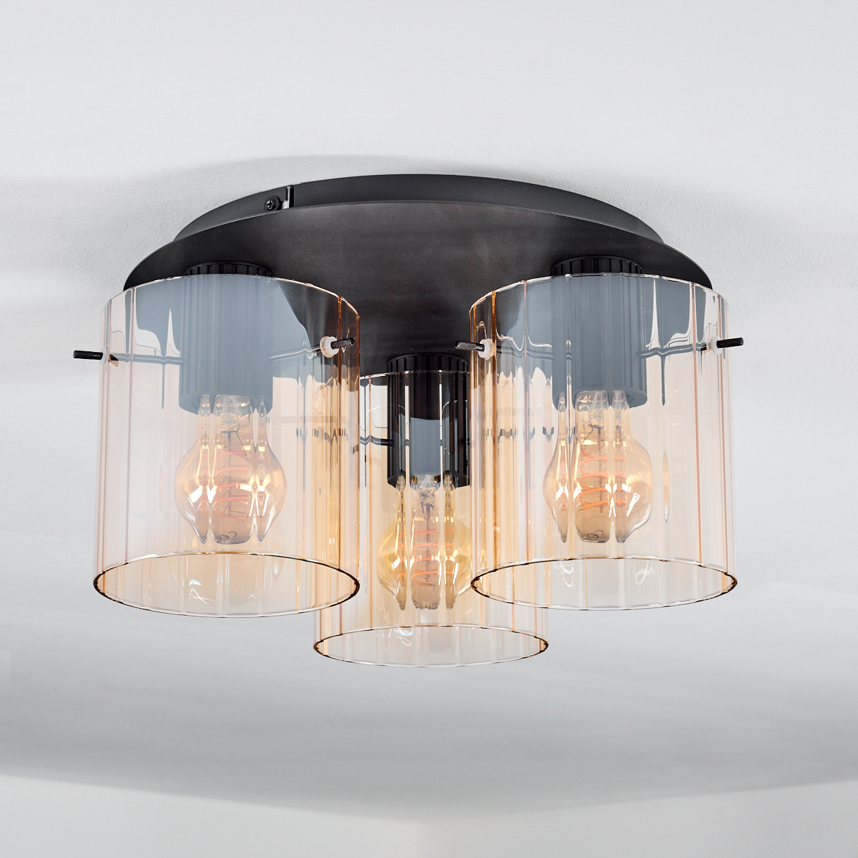 Eine moderne Deckenleuchte mit drei Lampen. Jede Lampe hat einen zylindrischen, bernsteinfarbenen Glasschirm und eine sichtbare Glühbirne.