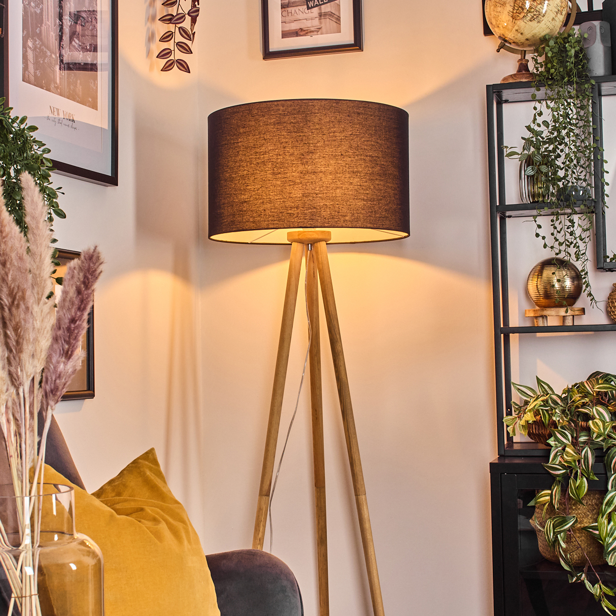 Ein Raum mit einer hölzernen Stativlampe, einem Regal mit Pflanzen, gerahmten Bildern und einer Couch mit gelben Kissen.