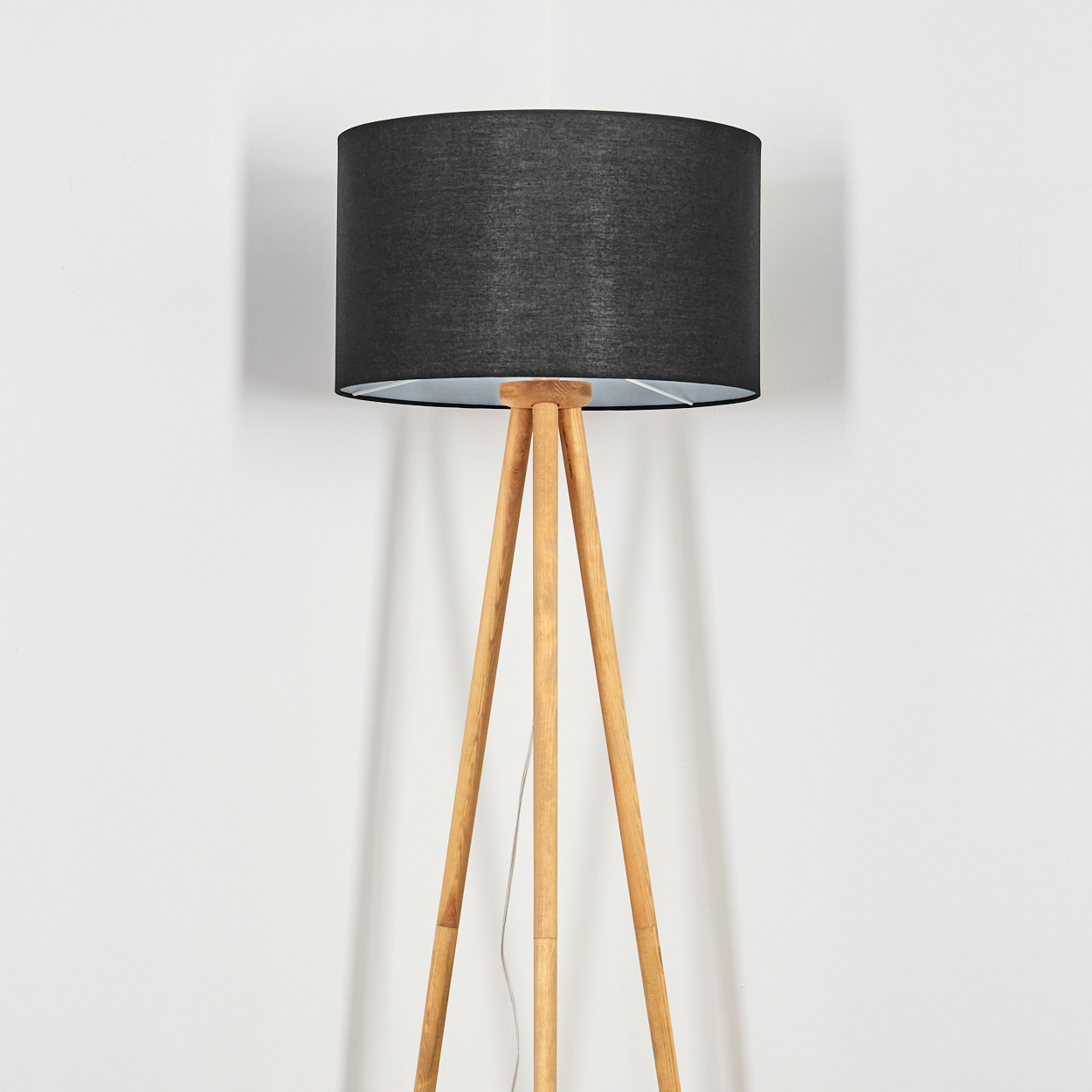 Eine Stehlampe mit schwarzem Lampenschirm und Holzstativbeinen steht vor einer weißen Wand. Ein Netzkabel ist sichtbar.