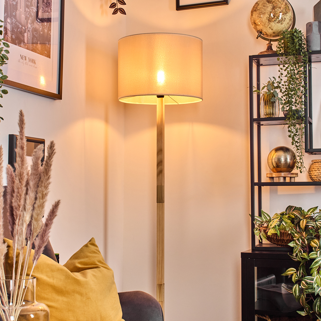 Eine beleuchtete Stehlampe steht in einer Ecke mit einem gelben Kissen und Pflanzen. Ein Regal hält einen Globus und Pflanzen. Ein New York Bild hängt.
