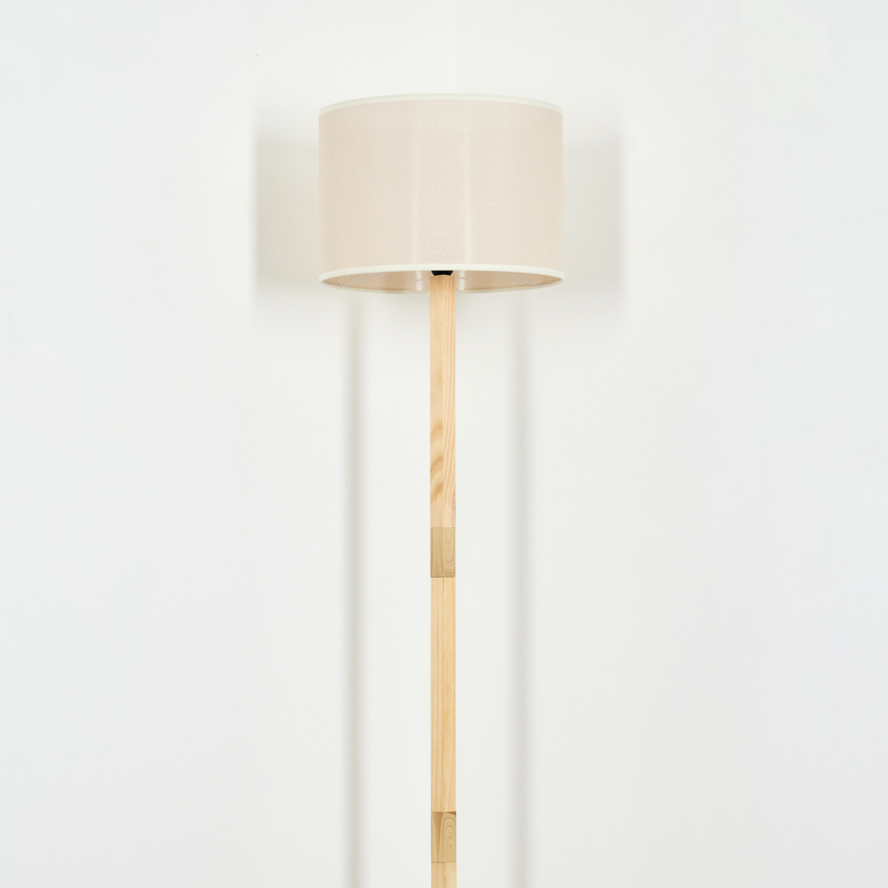 Ein beiger Lampenschirm steht auf einem Holzlampenständer vor einer weißen Wand. Die Lampe wirft einen Schatten.