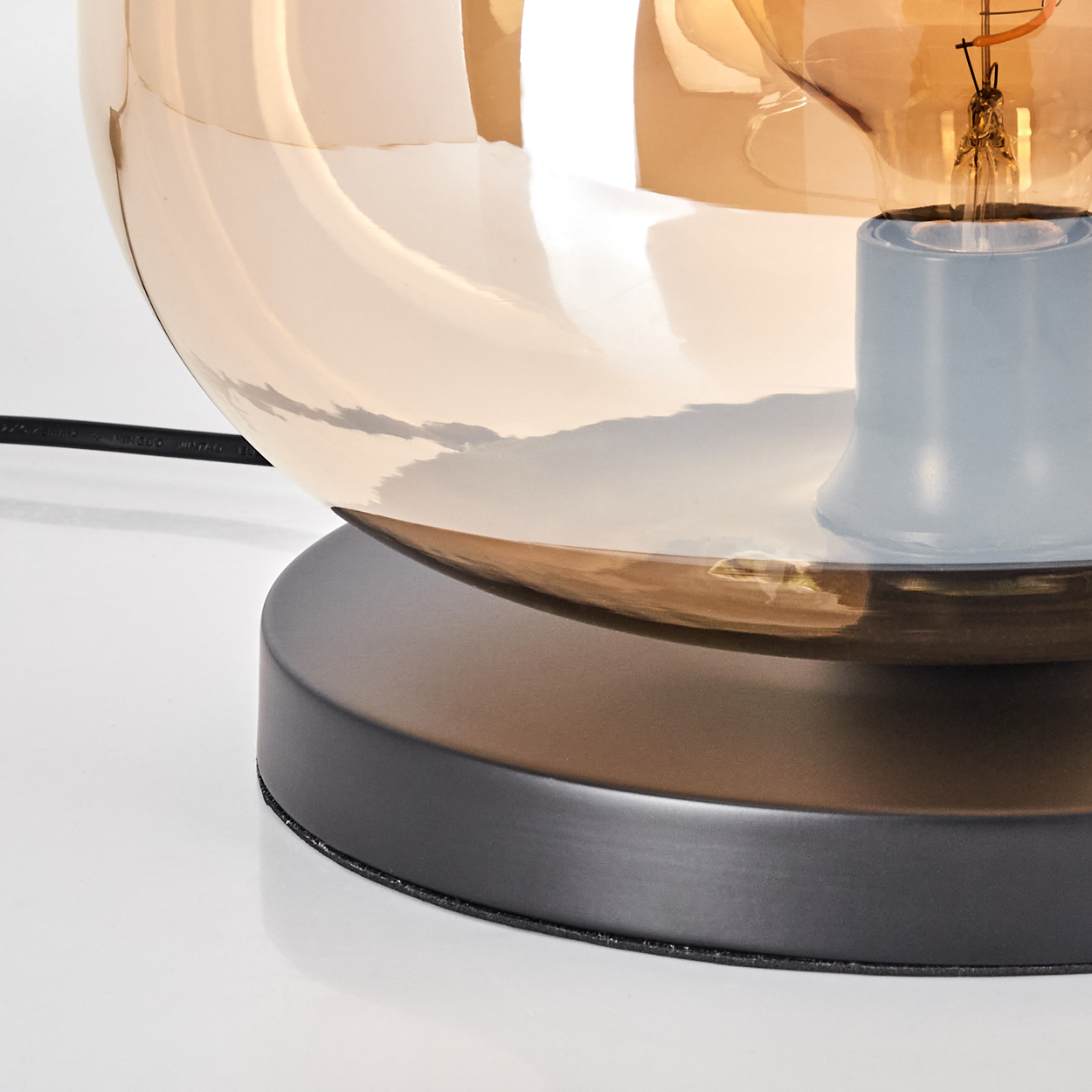 Eine moderne Lampe mit braunem Glasschirm und schwarzem Sockel. Eine Glühbirne ist im Schirm sichtbar. Der Sockel steht auf weißem Untergrund.