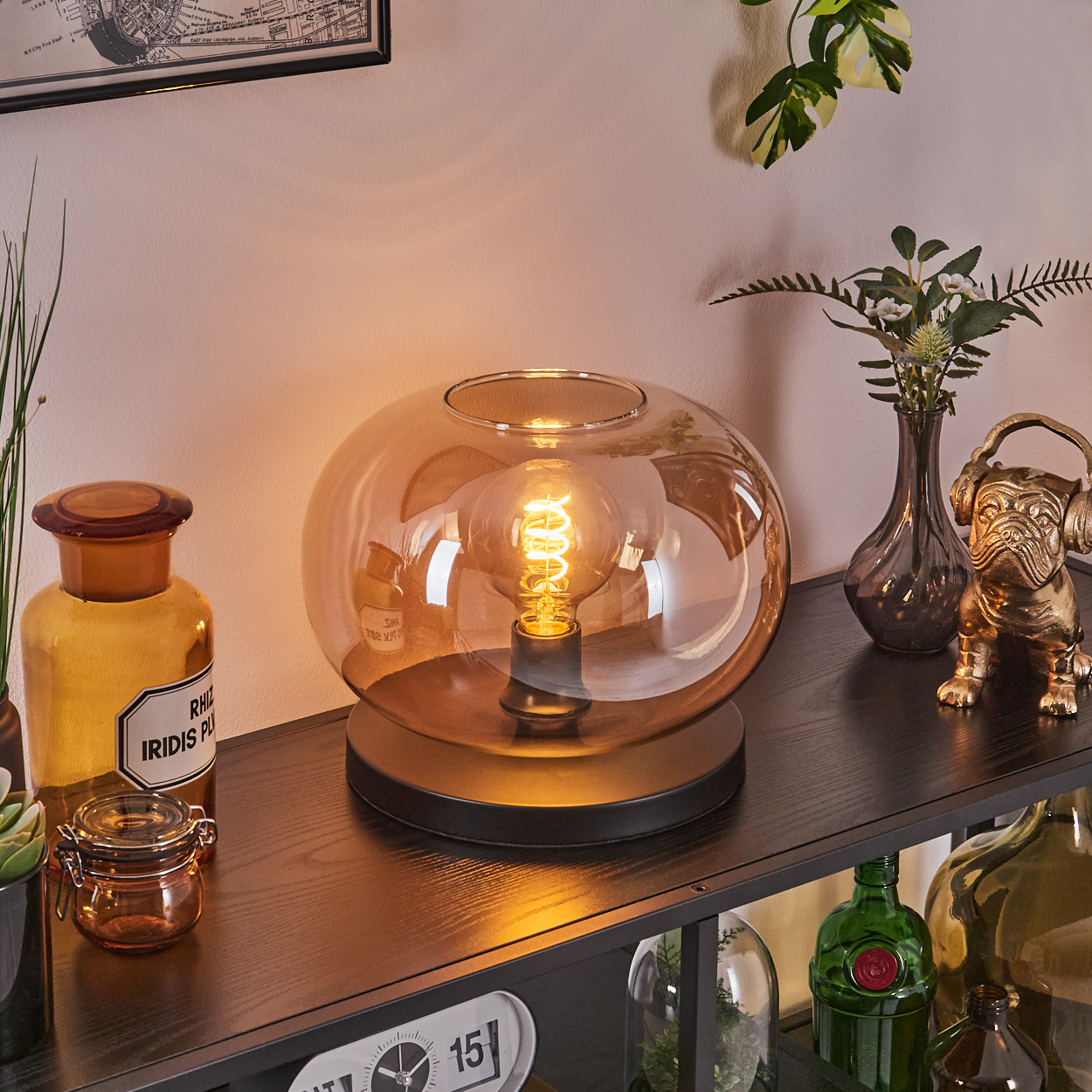 Ein dekoratives Regal mit einer Lampe, einer Bulldoggen-Statue und Glasbehältern. Die Lampe hat einen Glasschirm. Verschiedene grüne Pflanzen sind vorhanden.