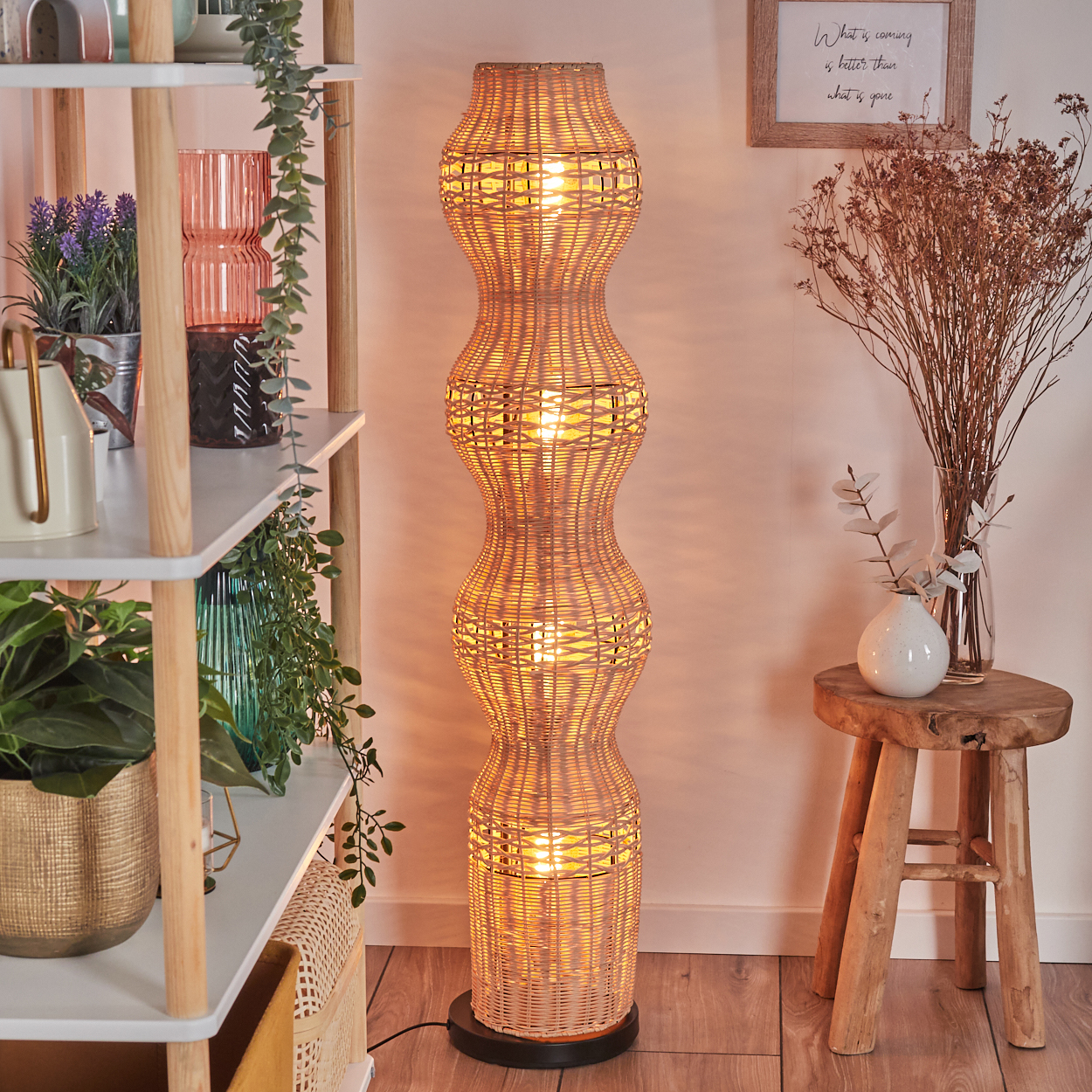 Eine gewebte Rattan-Stehlampe leuchtet, in der Nähe eines Holzschemels mit Vase und Trockenblumen. Regale halten Pflanzen und Dekorationsartikel.