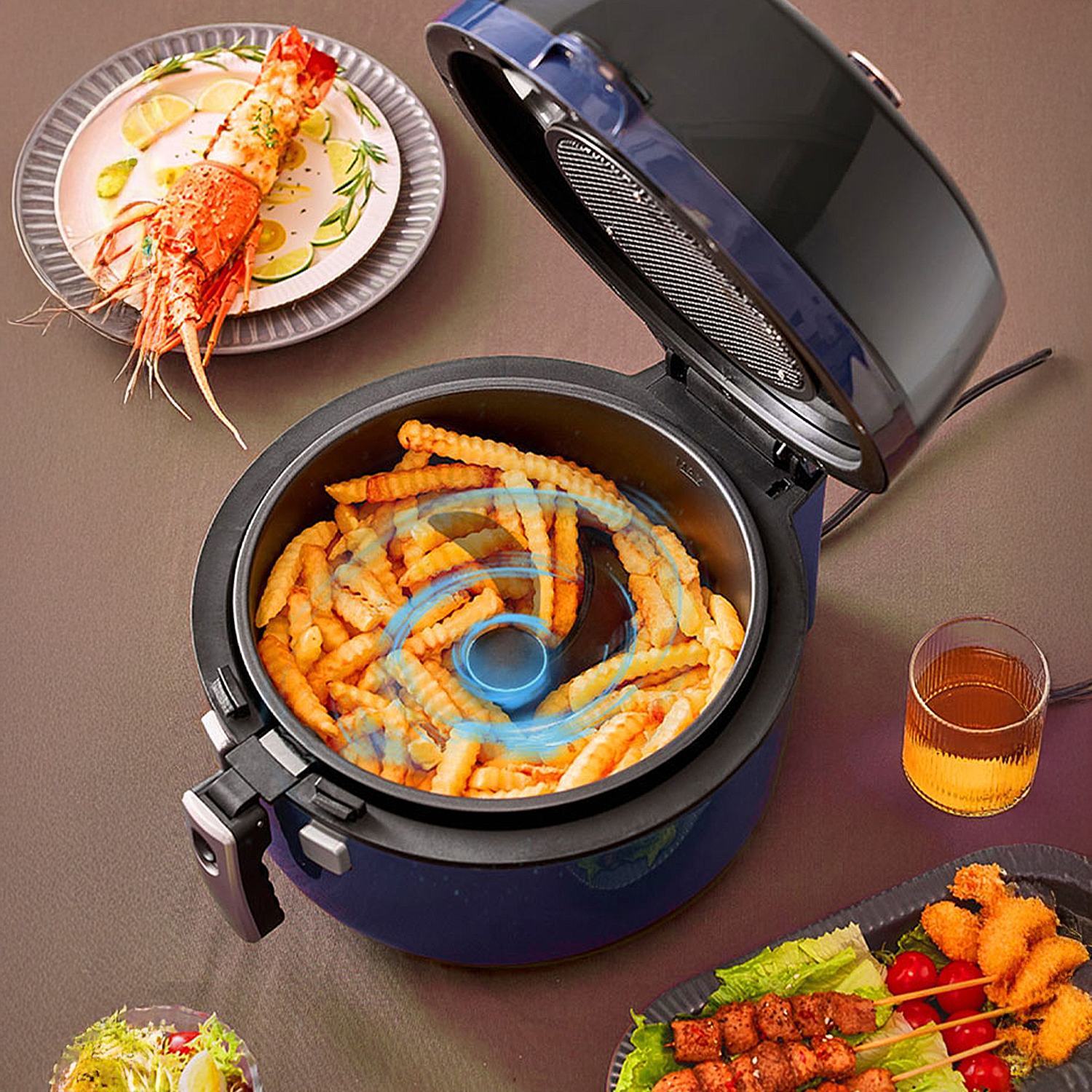 Una freidora de aire con papas fritas dentro, un plato con langosta y otros platos. La freidora está abierta y se ve un vaso con bebida.