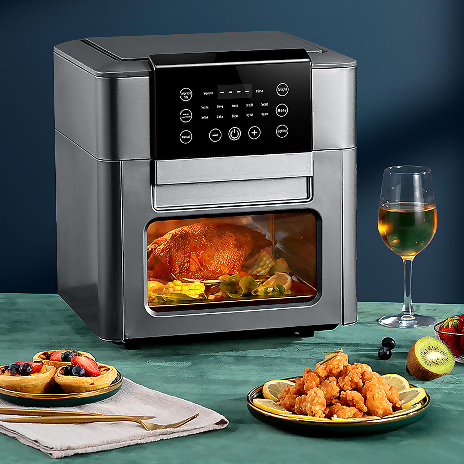 Una freidora de aire con pollo asado dentro, acompañada de tartaletas, comida frita, frutas y vino sobre una mesa verde.