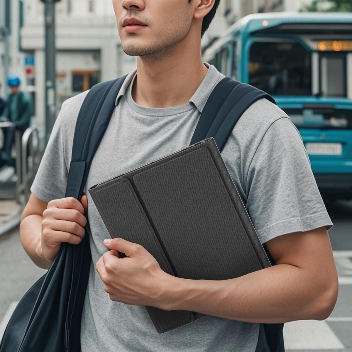 Ein Mann mit grauem T-Shirt und Rucksack hält eine schwarze Tablet-Hülle. Im Hintergrund ist ein Bus zu sehen.