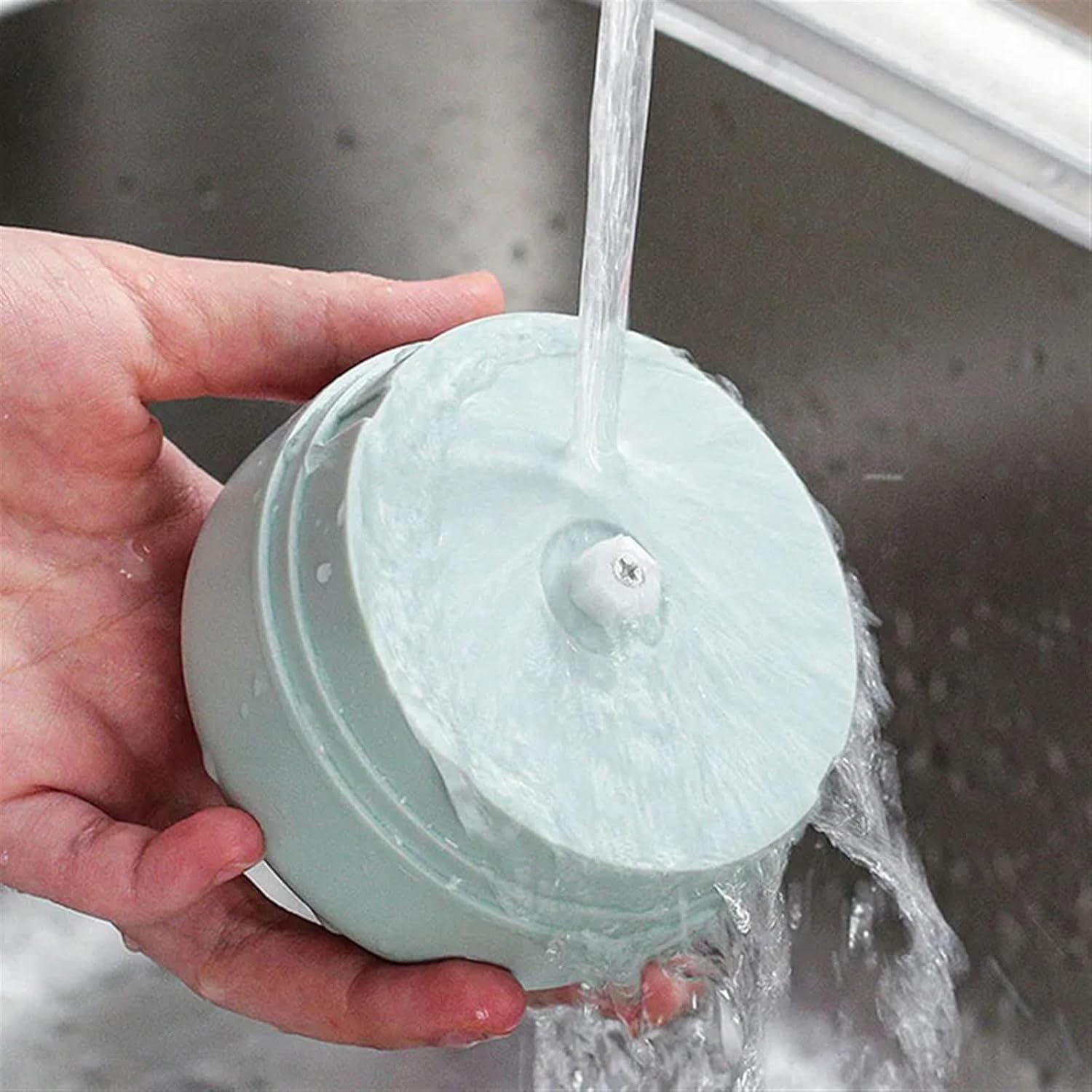 Una mano tiene un robot da cucina azzurro che viene lavato sotto l'acqua corrente in un lavandino in acciaio inossidabile.