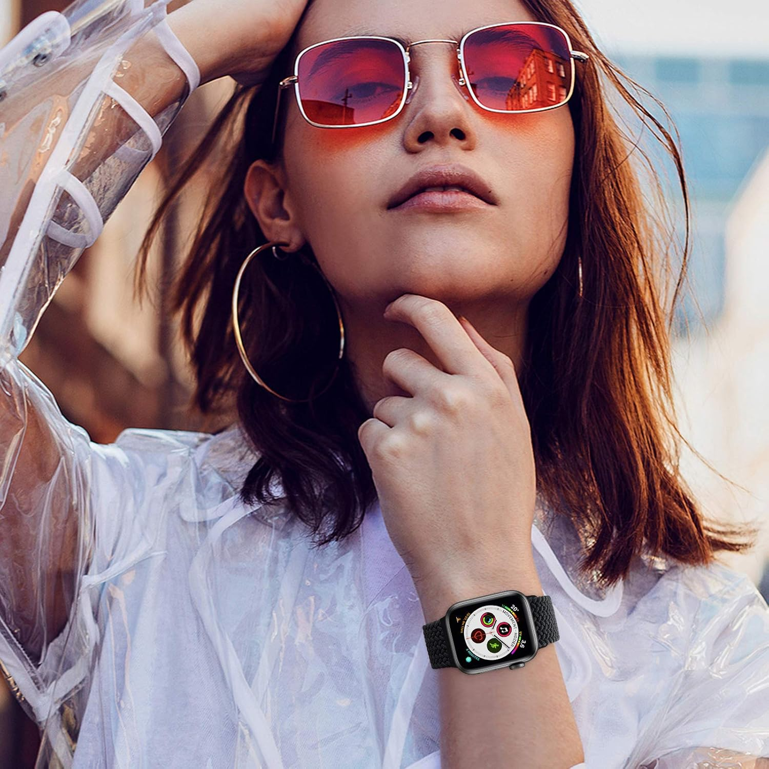 Frau mit roter Sonnenbrille, klarer Jacke und Smartwatch. Sie hat goldene Creolen und berührt ihr Gesicht.