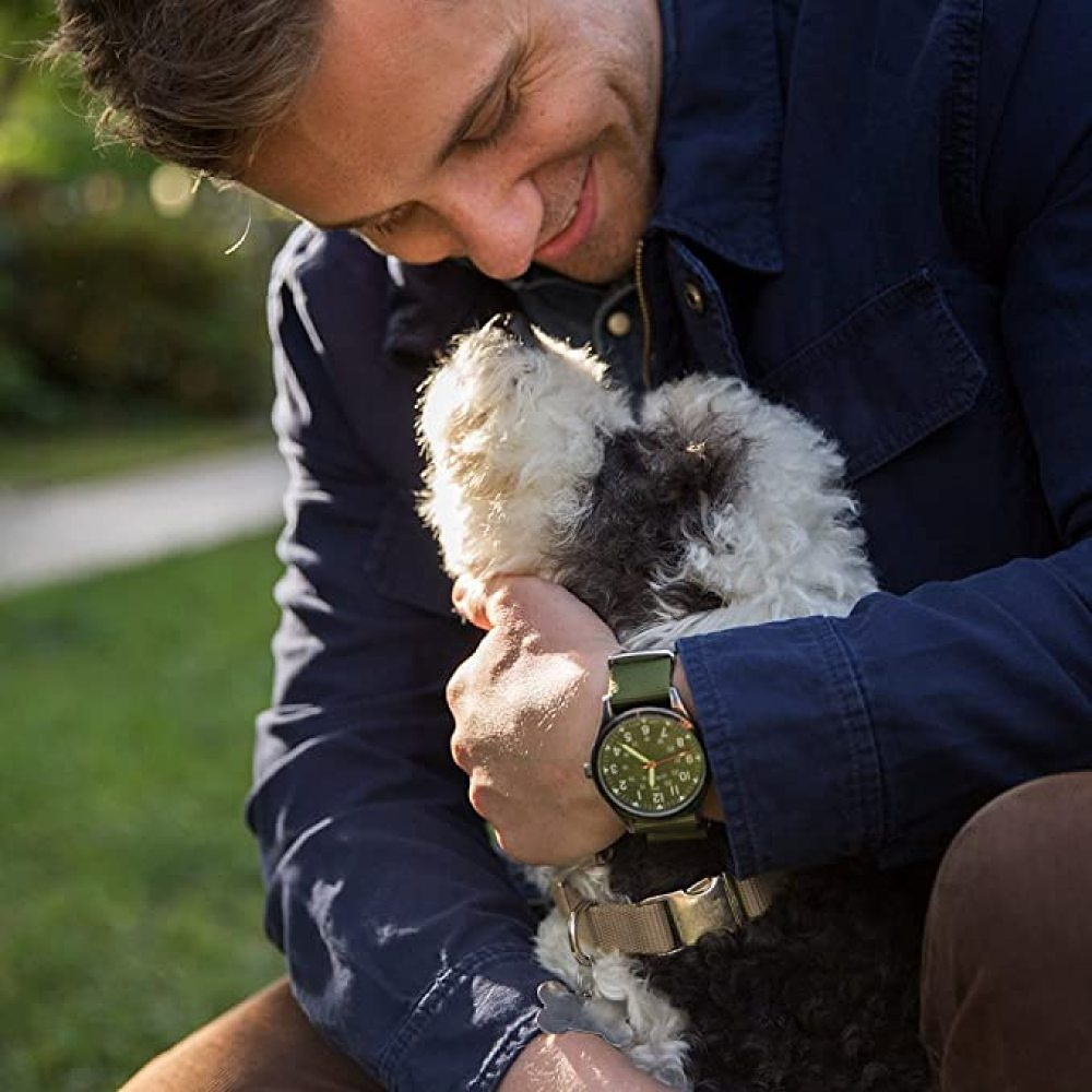Ein Mann lächelt, während er einen Hund hält. Der Hund ist schwarz und weiß. Der Mann trägt eine Uhr und eine blaue Jacke. Grünes Gras im Hintergrund.