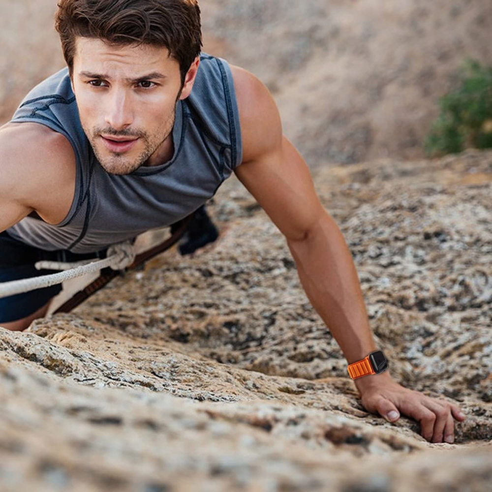 Ein Mann klettert eine Felswand hinauf. Er trägt ein graues Tanktop und eine orangefarbene Uhr. Ein Seil ist befestigt. Der Hintergrund ist verschwommene Felsen.