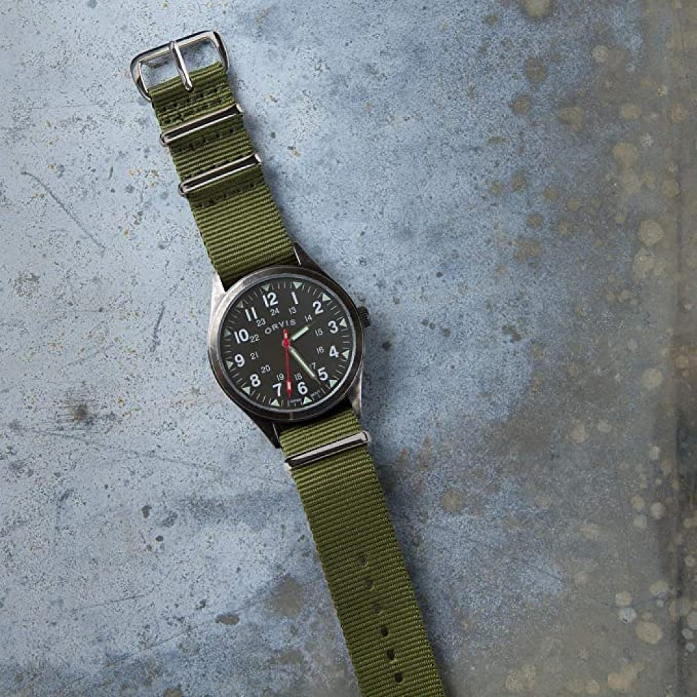 Eine Armbanduhr im Militärstil mit grünem Nylonarmband. Das Zifferblatt ist schwarz mit weißen Ziffern und Zeigern. Der Hintergrund ist grau.