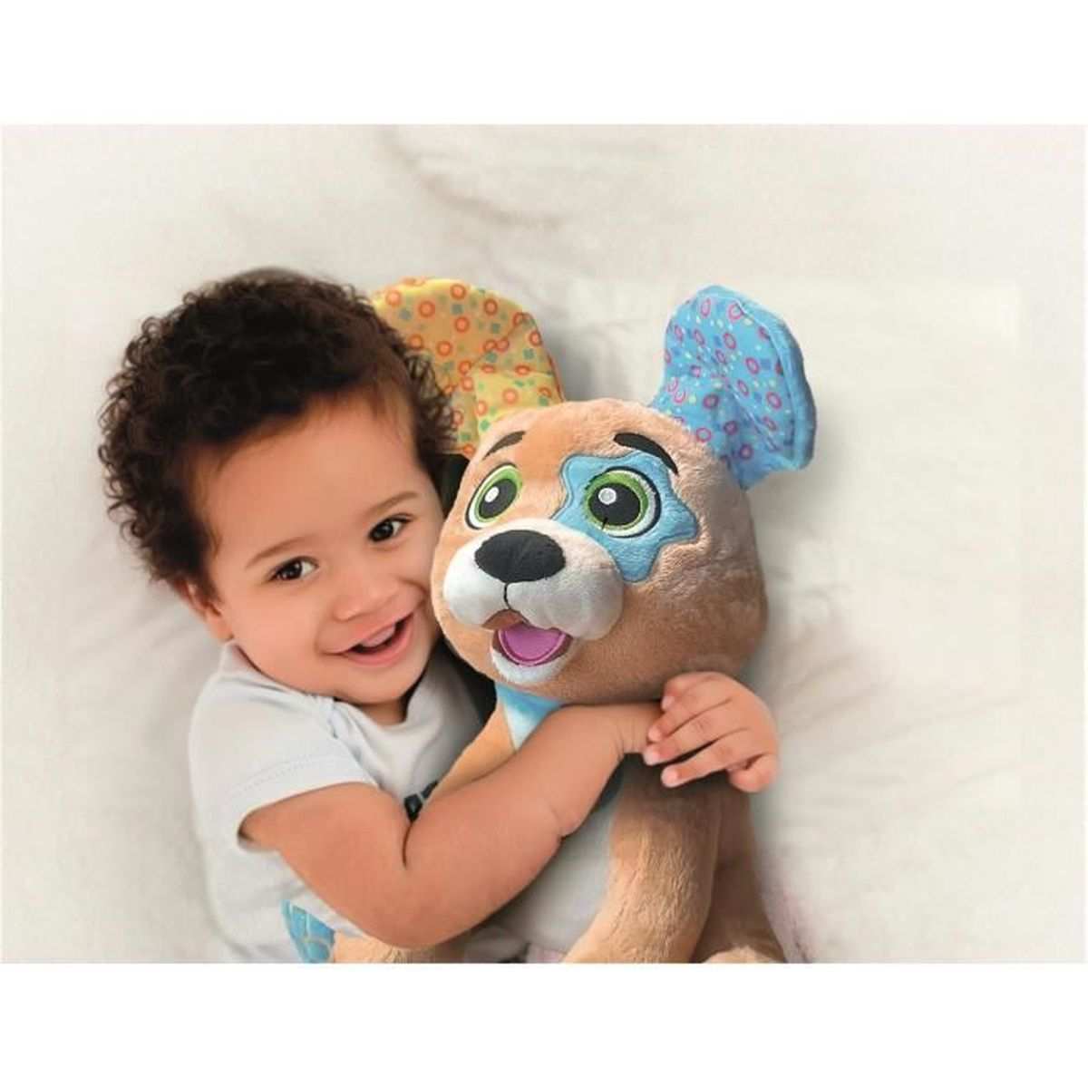 Ein lächelndes Baby umarmt einen Stoffhund. Der Hund ist braun mit blauen und grünen Akzenten. Das Baby trägt ein weißes Hemd.