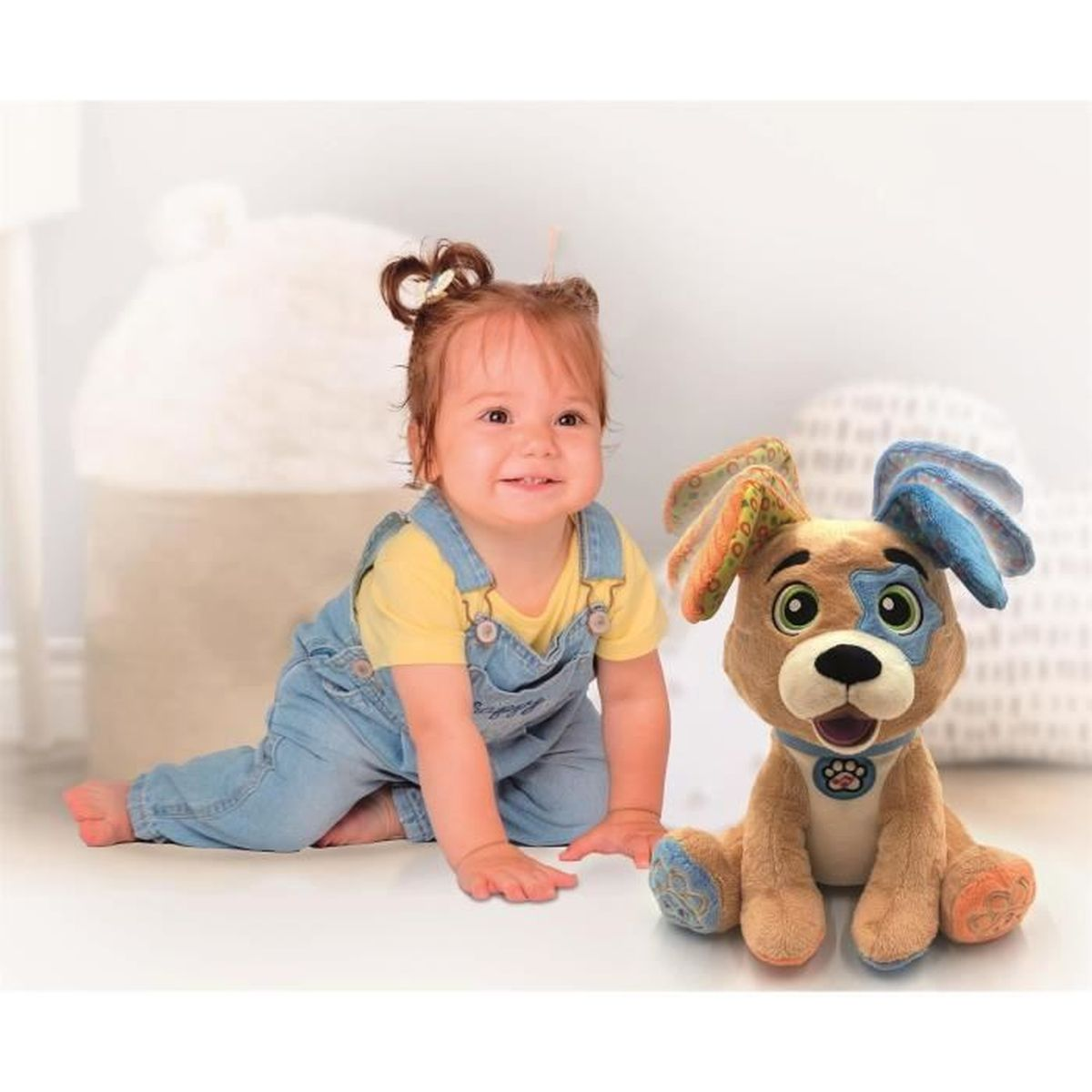 Ein lächelndes Baby in Latzhosen krabbelt in der Nähe eines Plüschhundespielzeugs. Der Hund ist braun mit blauen und grünen Akzenten. Im Hintergrund Kissen.