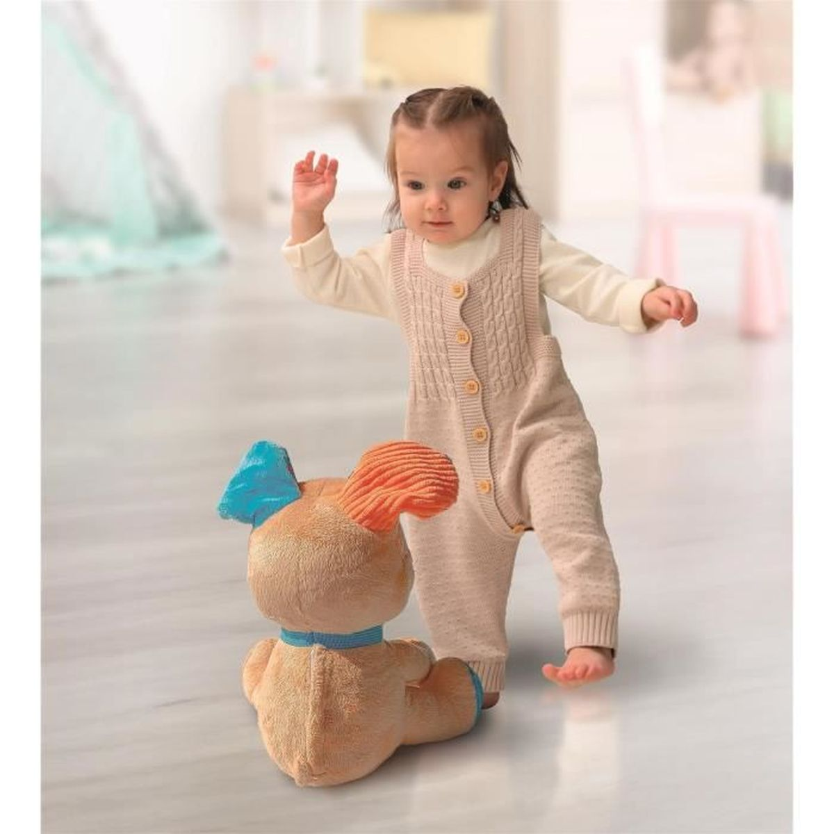 Ein Baby in einem gestrickten Overall steht in der Nähe eines Plüschhundespielzeugs. Das Baby greift danach. Das Spielzeug hat blaue und orange Ohren.