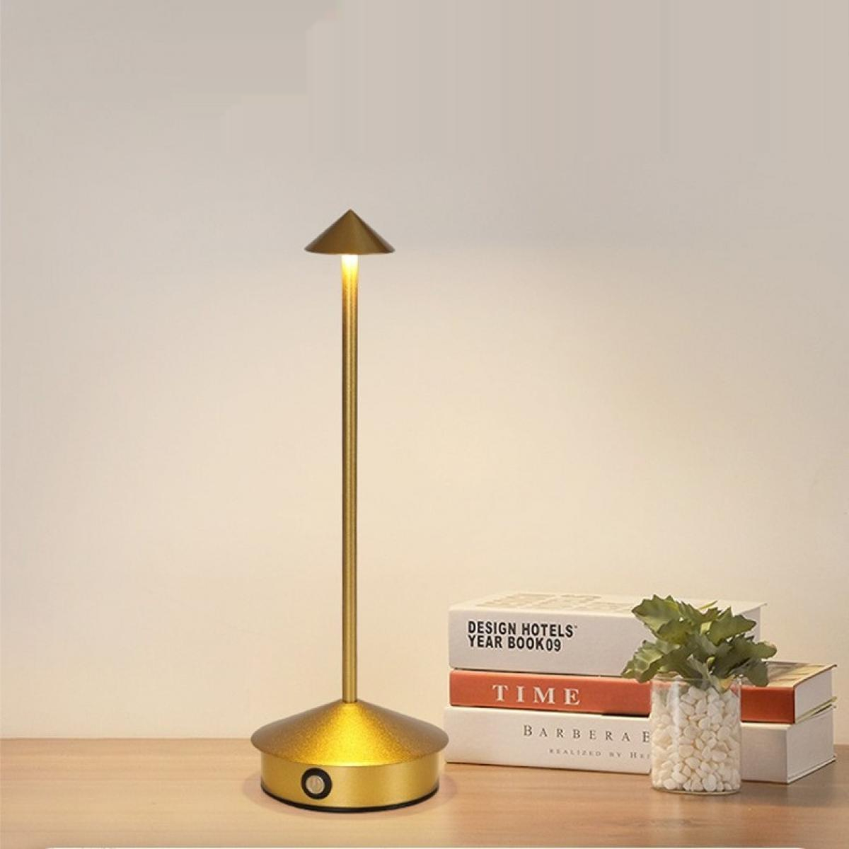 Eine goldene Lampe auf einem Holztisch, neben Büchern und einer kleinen Pflanze in einer Vase. Die Lampe ist beleuchtet.